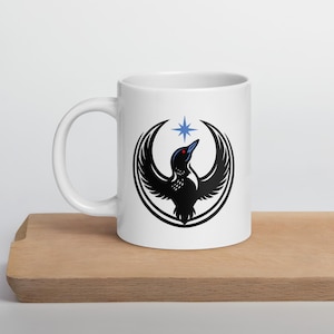 Peut inclure: Mug en céramique blanche avec un motif d'oiseau noir aux ailes déployées, un œil rouge et une étoile bleue au-dessus. L'oiseau est entouré d'un contour noir. Le mug est posé sur une surface en bois.