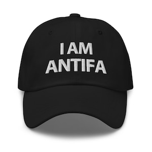 Op de afbeelding: Zwarte baseballcap met de geborduurde tekst "I AM ANTIFA" in het wit. De cap heeft een gebogen klep en een knoop aan de bovenkant. De hoed is gemaakt van een zacht materiaal.