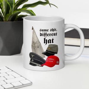 Peut inclure: Mug en céramique blanche avec l'inscription "Same shit, different hat" en police gothique noire. La tasse présente une cagoule blanche, une casquette militaire, une casquette noire et un chapeau rouge "Make America Great Again".