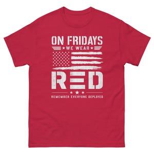 Peut inclure: Un t-shirt rouge avec un motif de drapeau américain vieilli et du texte blanc. Le texte indique "ON FRIDAYS WE WEAR RED" et "REMEMBER EVERYONE DEPLOYED". Le t-shirt a un col rond et des manches courtes.