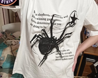 May include: White t-shirt with a black spider graphic and text. The graphic includes a star and lines resembling a web. Text on the shirt reads "When shepherds carrel the wol...Goas determine what you are going to be...Great wrks are ed by strength but by per eve rance Haste makes waste."