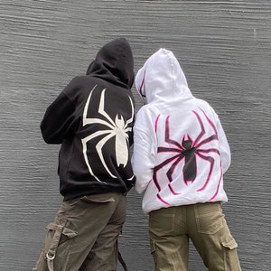 May include: Two hooded sweatshirts, one black and one white, each featuring a large spider graphic on the back. The black hoodie has a white spider design, while the white hoodie has a pink and black spider design. The people are wearing cargo pants and sneakers.