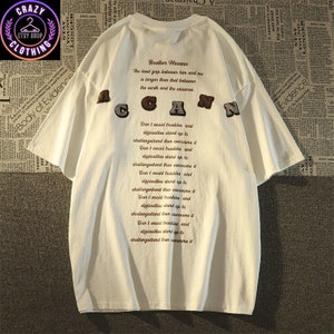 May include: White short-sleeve t-shirt with the word "ACGAN" in brown, gray, and black letters. The back of the shirt features text that reads "Brother Mecann" and a quote. The shirt has a relaxed fit.