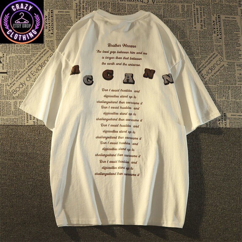 May include: White short-sleeve t-shirt with the word "ACGAN" in brown, gray, and black letters. The back of the shirt features text that reads "Brother Mecann" and a quote. The shirt has a relaxed fit.