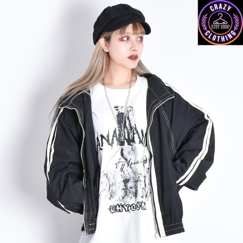 K&ouml;nnte beinhalten: Schwarze Jacke mit wei&szlig;en Streifen an den &Auml;rmeln, kombiniert mit einer schwarzen M&uuml;tze und einem wei&szlig;en Grafik-T-Shirt. Das T-Shirt zeigt ein schwarz-wei&szlig;es Design mit den Worten "ANARCHY" und "LHYOON". Eine silberne Halskette rundet den Look ab. Bild vom Crazy Clothing Etsy Shop.