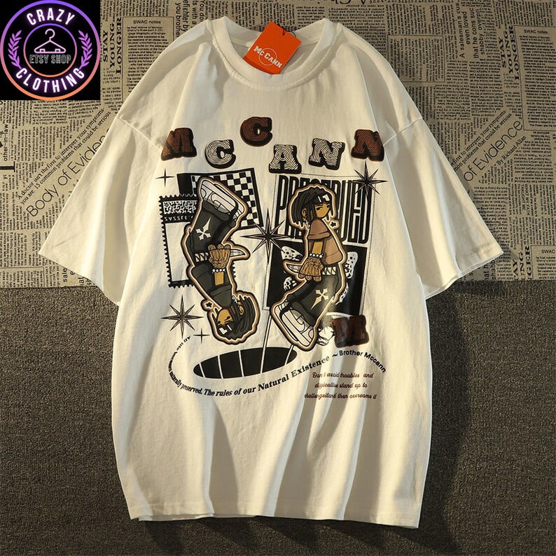 May include: White t-shirt with a graphic design featuring the text "McCann" in brown letters above a cartoon illustration. The illustration includes two figures and the text "Brother McCann". The shirt has short sleeves.