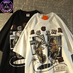 May include: Black and white t-shirts with a graphic design. The shirts feature the word "ACANA" in brown letters, along with an illustration of a person and the text "Brother Mccann". The shirts are from the Crazy Etsy Shop Clothing.