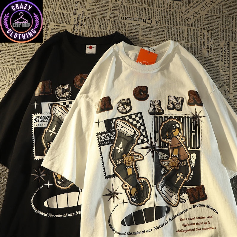 May include: Black and white t-shirts with a graphic design. The shirts feature the word "ACANA" in brown letters, along with an illustration of a person and the text "Brother Mccann". The shirts are from the Crazy Etsy Shop Clothing.