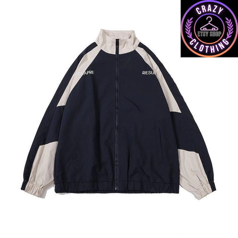 Op de afbeelding: Marineblauwe en beige trainingsjas met een lange ritssluiting. De jas heeft beige panelen op de schouders en mouwen, met de woorden "ETSUAPRE" en "RESU" op de borst gedrukt. Een "Crazy Clothing"-logo staat rechtsboven.
