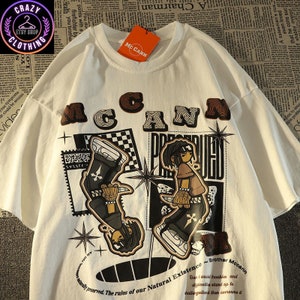 May include: White t-shirt with a graphic design featuring the word "McCann" in brown letters above two cartoon figures. The shirt has additional text and starburst designs. A tag reads "McCann".