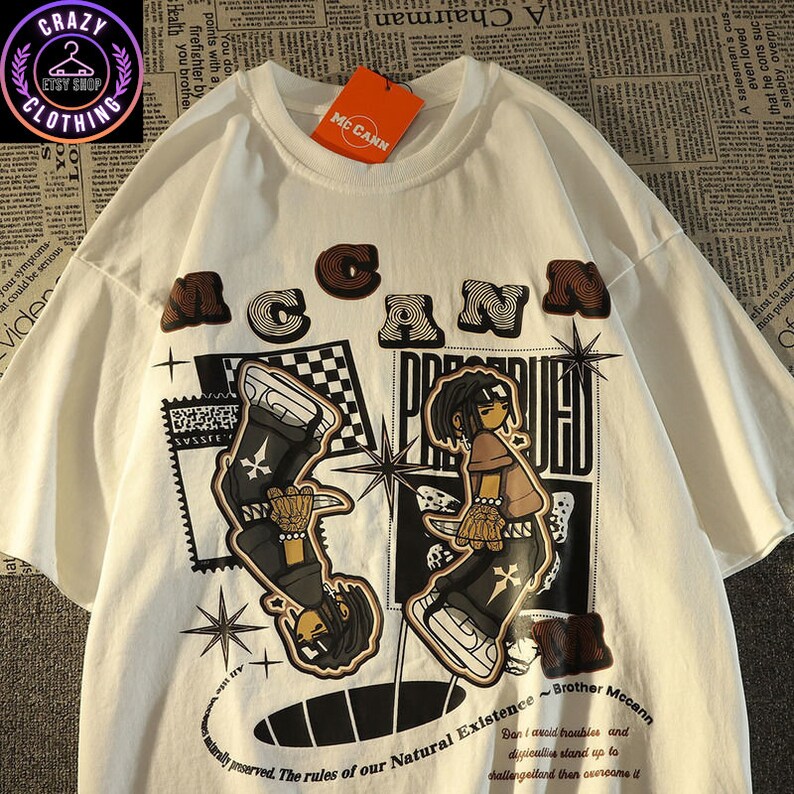 May include: White t-shirt with a graphic design featuring the word "McCann" in brown letters above two cartoon figures. The shirt has additional text and starburst designs. A tag reads "McCann".