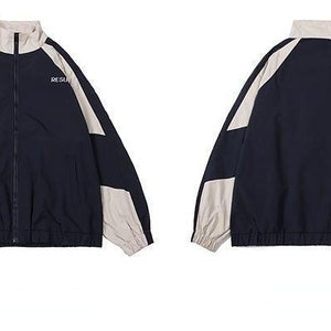 Op de afbeelding: Marineblauwe en beige trainingsjas met een volledige ritssluiting en een opstaande kraag. De jas heeft beige panelen op de schouders en mouwen, met de woorden "RESURE" en "RESURS" op de borst gedrukt. De jas heeft elastische manchetten en zoom.