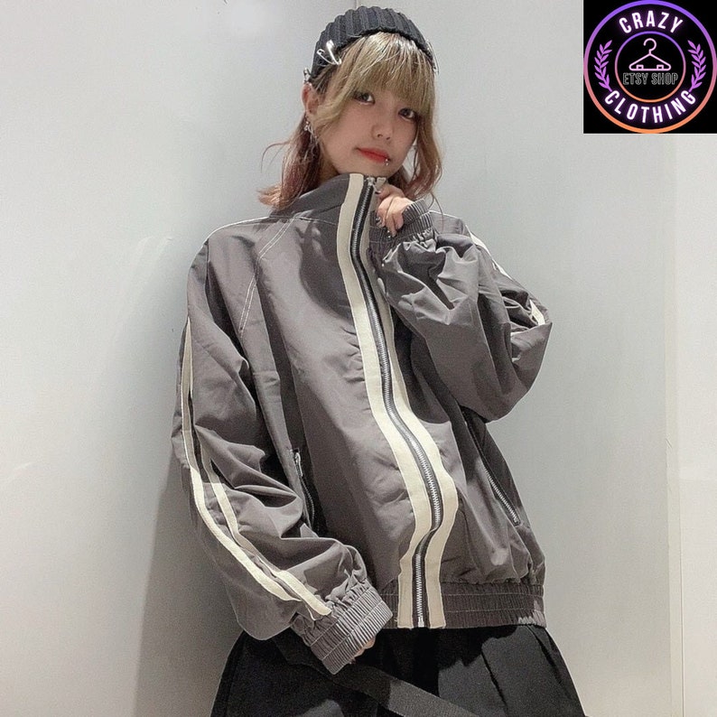 K&ouml;nnte beinhalten: Graue Jacke mit Rei&szlig;verschluss und wei&szlig;en Streifen an &Auml;rmeln und Vorderseite. Die Jacke hat einen hohen Kragen und gerippte B&uuml;ndchen und Saum. Das Logo des Crazy Clothing Etsy Shops ist oben rechts zu sehen.
