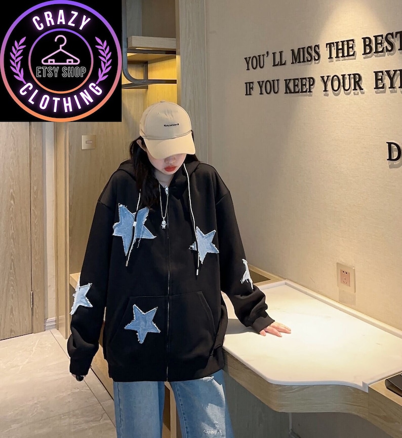 May include: Black zip-up hoodie with light blue star patches. The hoodie has a drawstring hood and a front zipper. The image also shows a beige baseball cap and blue jeans. The Etsy shop logo is in the upper left corner.