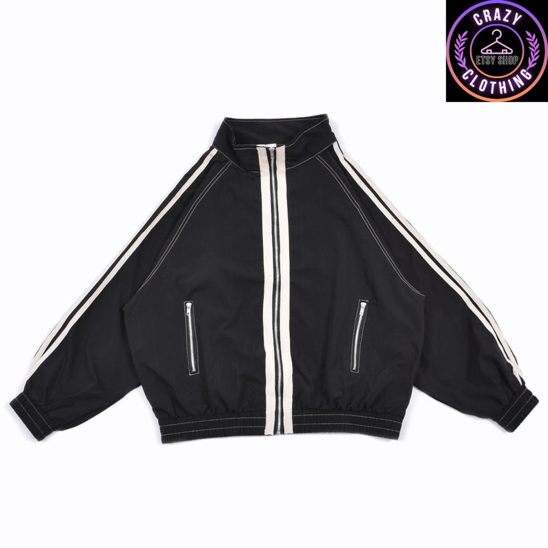 K&ouml;nnte beinhalten: Schwarze Trainingsjacke mit wei&szlig;en Streifen an &Auml;rmeln und Vorderseite. Die Jacke hat einen durchgehenden Rei&szlig;verschluss, zwei Seitentaschen sowie einen gerippten Saum und B&uuml;ndchen. Sie hat einen hohen Kragen und wei&szlig;e Nahtdetails.