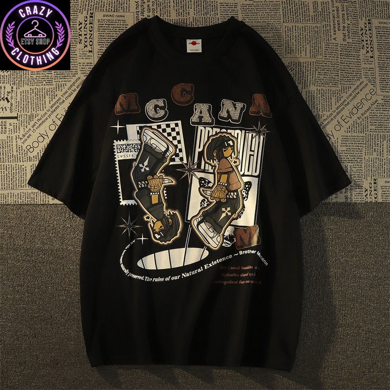 May include: Black graphic t-shirt featuring a stylized illustration with the text "MACANA PROUD". The design includes two figures, one inverted, with a checkerboard pattern and star accents. The shirt also has additional text.