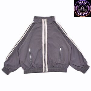 K&ouml;nnte beinhalten: Graue Trainingsjacke mit hohem Kragen, durchgehendem Rei&szlig;verschluss und zwei Vordertaschen. Die Jacke hat wei&szlig;e Streifen an den &Auml;rmeln und einen wei&szlig;en Streifen in der Mitte. Das Logo "CRAZY CLOTHING" ist sichtbar.