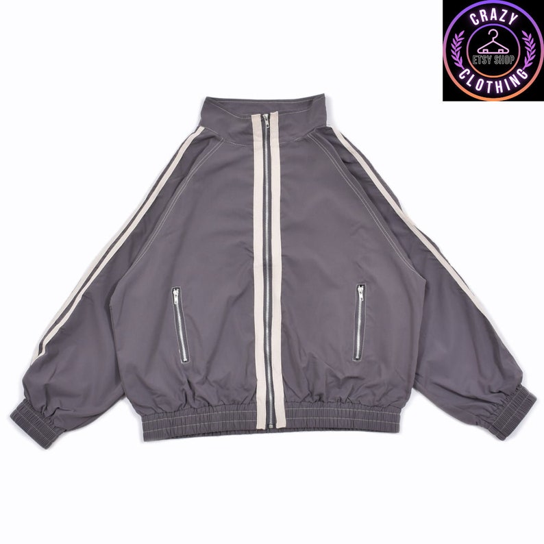 K&ouml;nnte beinhalten: Graue Trainingsjacke mit hohem Kragen, durchgehendem Rei&szlig;verschluss und zwei Vordertaschen. Die Jacke hat wei&szlig;e Streifen an den &Auml;rmeln und einen wei&szlig;en Streifen in der Mitte. Das Logo "CRAZY CLOTHING" ist sichtbar.