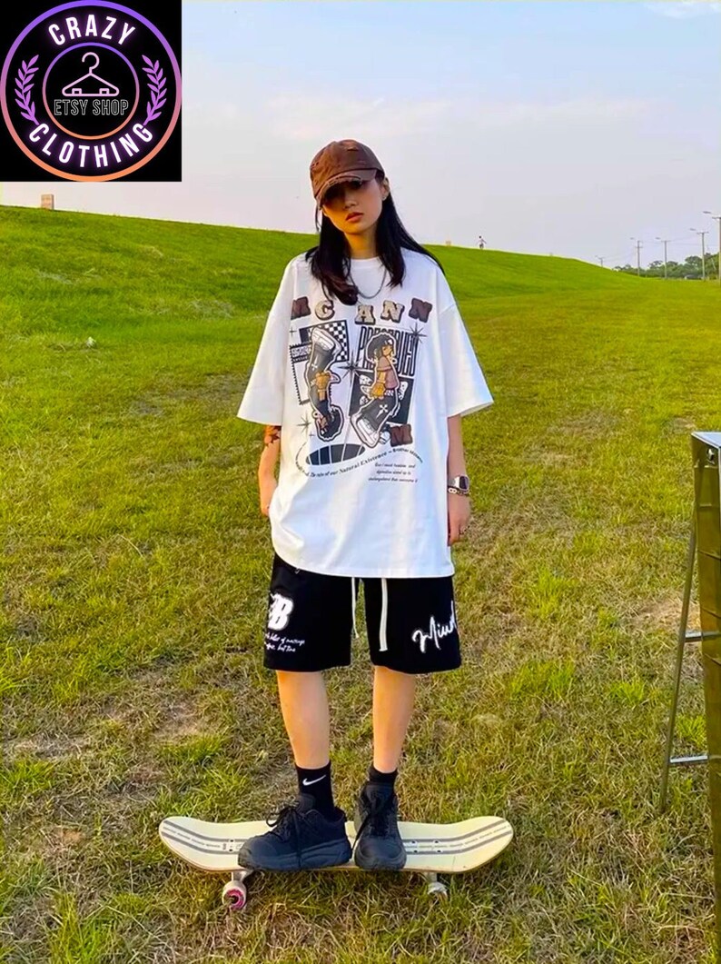 May include: A person wearing a white graphic tee and black shorts stands on a skateboard. The tee features a cartoon design with the word "CANN" and the shorts have white drawstrings and a white graphic. The person is wearing a brown cap and black shoes.