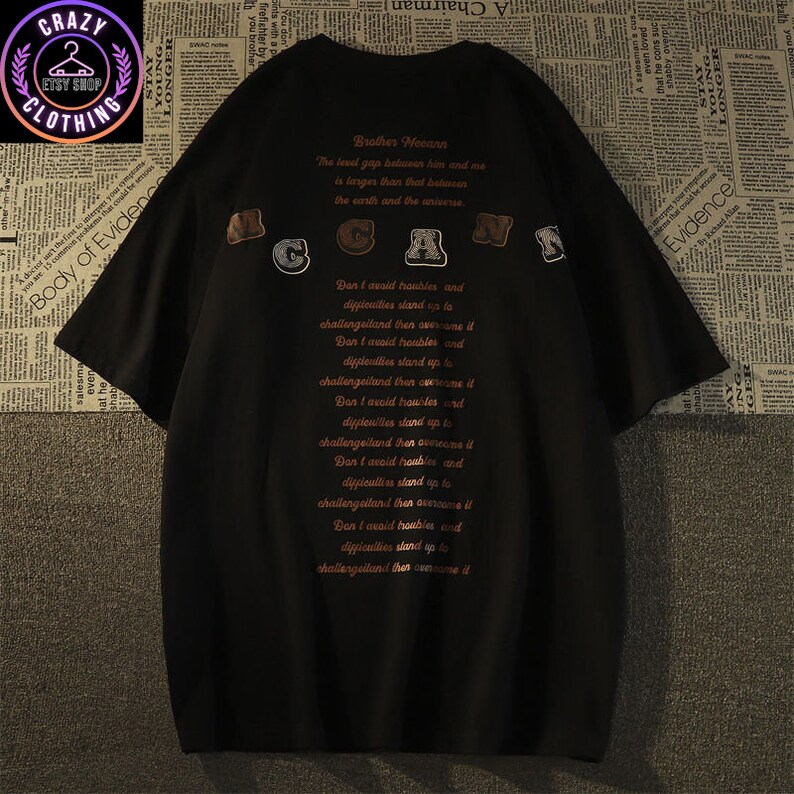 May include: Black oversized t-shirt with the text "Brother McCann" and a list of inspirational phrases in a reddish-brown font. The shirt features the word "McCann" in a decorative font across the top.
