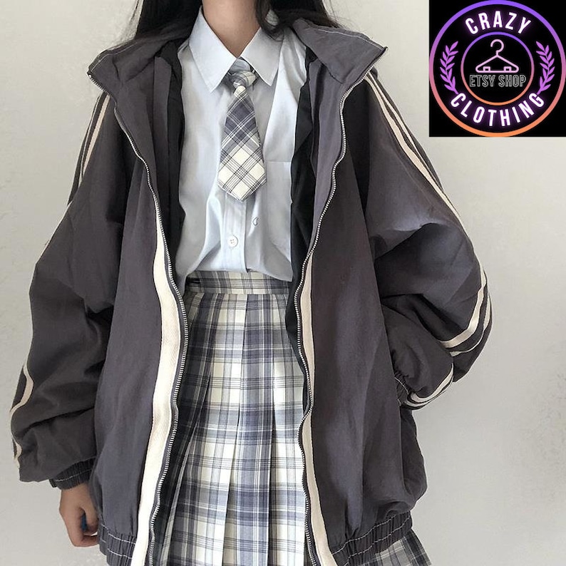 K&ouml;nnte beinhalten: Dunkelgraue Jacke mit Rei&szlig;verschluss und wei&szlig;en Streifen an den &Auml;rmeln, getragen &uuml;ber einem hellblauen Hemd, einem karierten Rock und einer Krawatte. Die Jacke hat eine Kapuze und einen wei&szlig;en Rei&szlig;verschluss. Das Bild enth&auml;lt ein Logo "CRAZY CLOTHING".