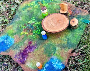 May include: A felt play mat with a green, brown, and blue landscape. There are three wooden figures and a wooden table and chair on the mat.
