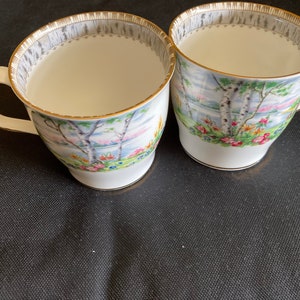 May include: Two white teacups with a gold rim and a floral pattern featuring a lake scene with birch trees. The cups are on a black surface.