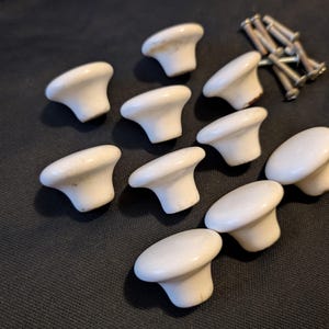 May include: A collection of nine white ceramic cabinet knobs with a smooth, rounded top and a tapered base, accompanied by a small pile of silver-coloured metal mounting screws. The knobs are arranged in a scattered pattern across the frame.