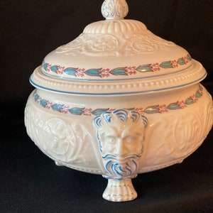 May include: A white ceramic soup tureen with a lid, featuring blue and red floral accents. The tureen has a sculpted face design and rests on three feet. The lid is topped with a decorative finial.