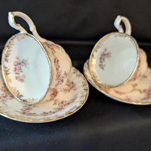 May include: Two white porcelain teacups and matching saucers featuring a delicate pink floral pattern and gold gilded scalloped edges. Each cup has a curved handle and a fluted design, with the floral motif repeated on the interior and exterior.