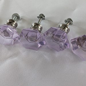 May include: Four faceted, lavender-coloured glass cabinet knobs with silver-toned metal hardware. The knobs have a geometric design and are arranged on a white surface. These decorative knobs are suitable for furniture or cabinet upgrades.