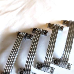 May include: Four vintage silver cabinet handles. Each handle features a curved, ribbed design and is attached to a rectangular base with screw holes. The handles are arranged on a white surface.