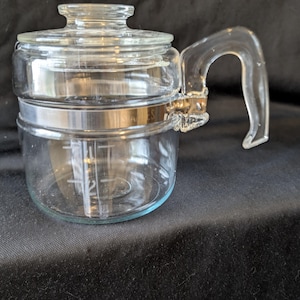 May include: A clear glass coffee pot with a lid and handle. The pot has a metal band around the middle and measurement markings on the side. The handle is curved and attached to the side of the pot.