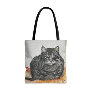 May include: A tote bag featuring a watercolor painting of a gray tabby cat. The cat is depicted in a relaxed pose, with green eyes and a detailed fur pattern. The bag has black handles and a neutral background.