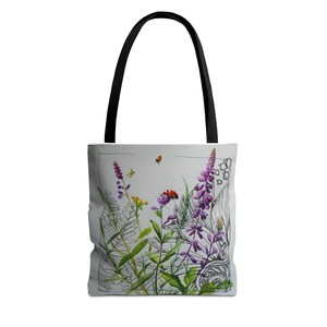 Può includere: Una borsa tote bianca con manici neri presenta un design floreale colorato. Il design include fiori viola, foglie verdi, una coccinella rossa e farfalle gialle. Lo sfondo è grigio chiaro.