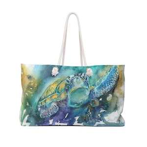 Può includere: Una grande borsa tote con un dipinto ad acquerello di una tartaruga marina. La tartaruga è raffigurata in tonalità di blu, verde e giallo, con un manico in corda bianca. La borsa ha una forma rettangolare ed è probabilmente in tessuto.