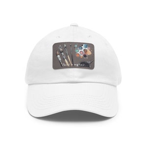 Puede incluir: Gorra de béisbol blanca con un parche marrón que muestra pinceles, una paleta de pintura y el texto "Paint in my hair..."