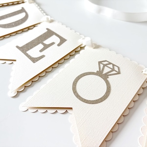 May include: Cream-colored banner with scalloped edges, featuring gray letters and a diamond ring illustration. The banner is strung with a white ribbon, perfect for a wedding or engagement celebration.