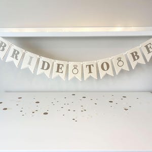May include: A banner that reads "BRIDE TO BE" in gray letters, with a ring graphic, hangs against a white background. The banner is made of off-white pennant flags with scalloped edges. The banner is a wedding decoration.