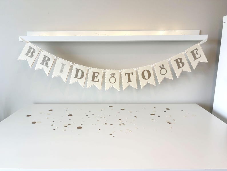 May include: A banner that reads "BRIDE TO BE" in gray letters, with a ring graphic, hangs against a white background. The banner is made of off-white pennant flags with scalloped edges. The banner is a wedding decoration.