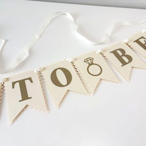 May include: A banner with the words "TO BE" in gold lettering, with a diamond ring graphic on the first and third flags. The flags are cream-colored with scalloped edges and are strung on a white ribbon.