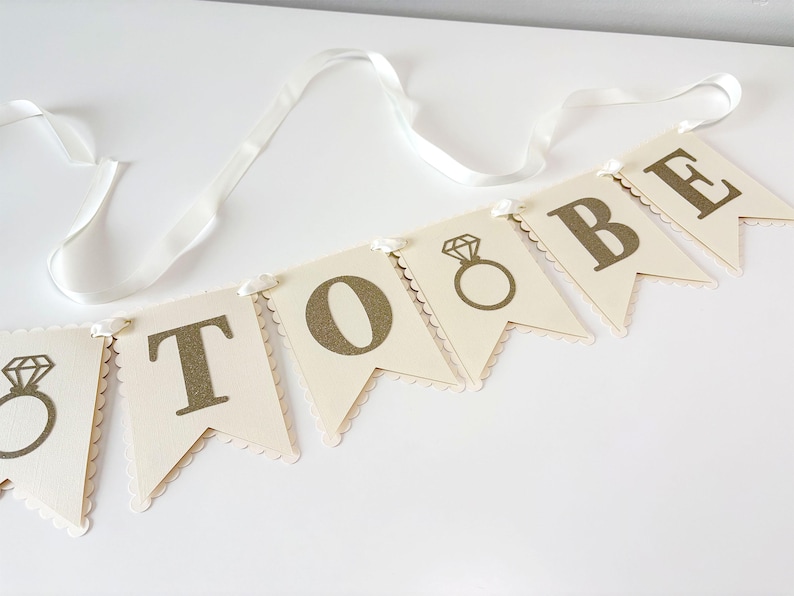 May include: A banner with the words "TO BE" in gold lettering, with a diamond ring graphic on the first and third flags. The flags are cream-colored with scalloped edges and are strung on a white ribbon.