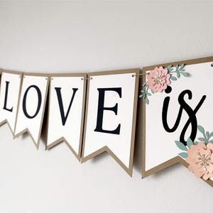 Op de afbeelding: Een decoratieve slinger met witte wimpels met bruine randen aan een dun koord. De letters L, O, V, E en het woord is zijn in het zwart gedrukt. Roze papieren bloemen met groene bladeren zijn bevestigd aan de eerste en laatste wimpels.