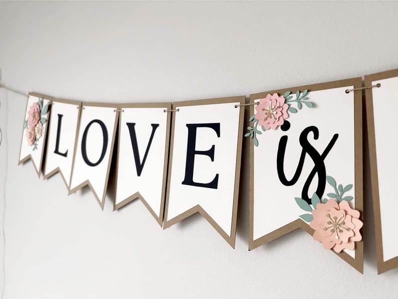 Op de afbeelding: Een decoratieve slinger met witte wimpels met bruine randen aan een dun koord. De letters L, O, V, E en het woord is zijn in het zwart gedrukt. Roze papieren bloemen met groene bladeren zijn bevestigd aan de eerste en laatste wimpels.