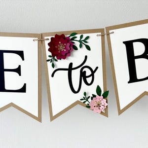 Op de afbeelding: Een decoratieve slinger bestaande uit witte vlaggetjes met bruine randen aan een touw. De vlaggetjes tonen de zwarte letters E en B, met het woord to in zwarte schrijfletters. Een donkerrode bloem en een lichtroze bloem met groene bladeren zijn bevestigd.