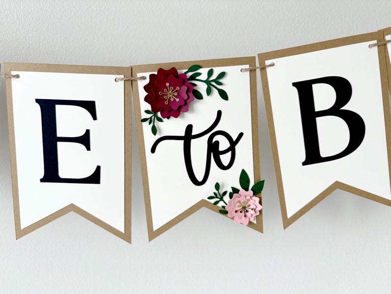 Op de afbeelding: Een decoratieve slinger bestaande uit witte vlaggetjes met bruine randen aan een touw. De vlaggetjes tonen de zwarte letters E en B, met het woord to in zwarte schrijfletters. Een donkerrode bloem en een lichtroze bloem met groene bladeren zijn bevestigd.
