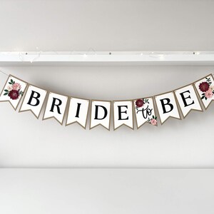 Op de afbeelding: Een decoratieve slinger bestaande uit witte vlaggetjes met zwarte letters die de tekst BRIDE TO BE vormen. De buitenste vlaggetjes zijn versierd met bloemillustraties in bordeauxrood, roze en groen. De slinger hangt aan een dun koord.