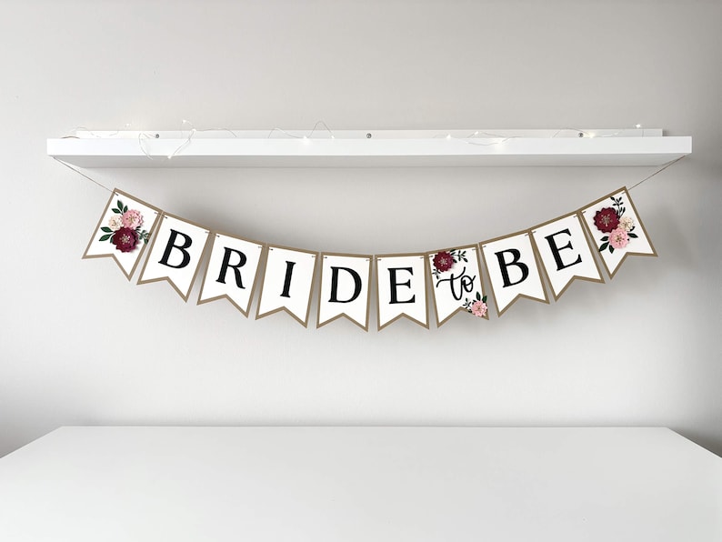 Op de afbeelding: Een decoratieve slinger bestaande uit witte vlaggetjes met zwarte letters die de tekst BRIDE TO BE vormen. De buitenste vlaggetjes zijn versierd met bloemillustraties in bordeauxrood, roze en groen. De slinger hangt aan een dun koord.