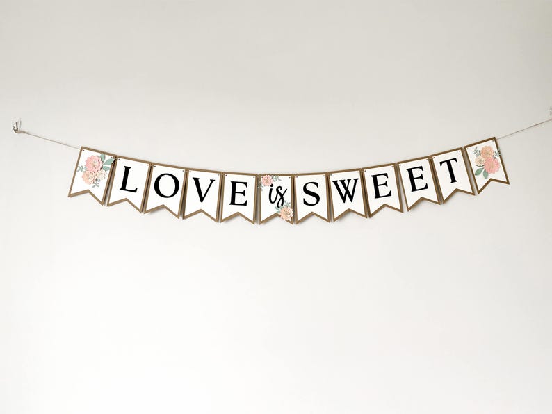 Op de afbeelding: Een decoratieve slinger bestaande uit individuele witte vlaggetjes met bruine randen, die de tekst LOVE IS SWEET vormen in zwarte hoofdletters. De twee buitenste vlaggetjes en het middelste vlaggetje zijn versierd met illustraties van roze bloemen.