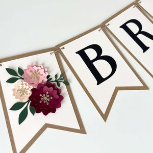 Op de afbeelding: Een decoratieve slinger bestaande uit witte kartonnen vlaggetjes met bruine randen aan een touwtje. E&eacute;n vlaggetje is versierd met driedimensionale papieren bloemen in roze, bordeaux en groen. Andere vlaggetjes bevatten grote zwarte hoofdletters.